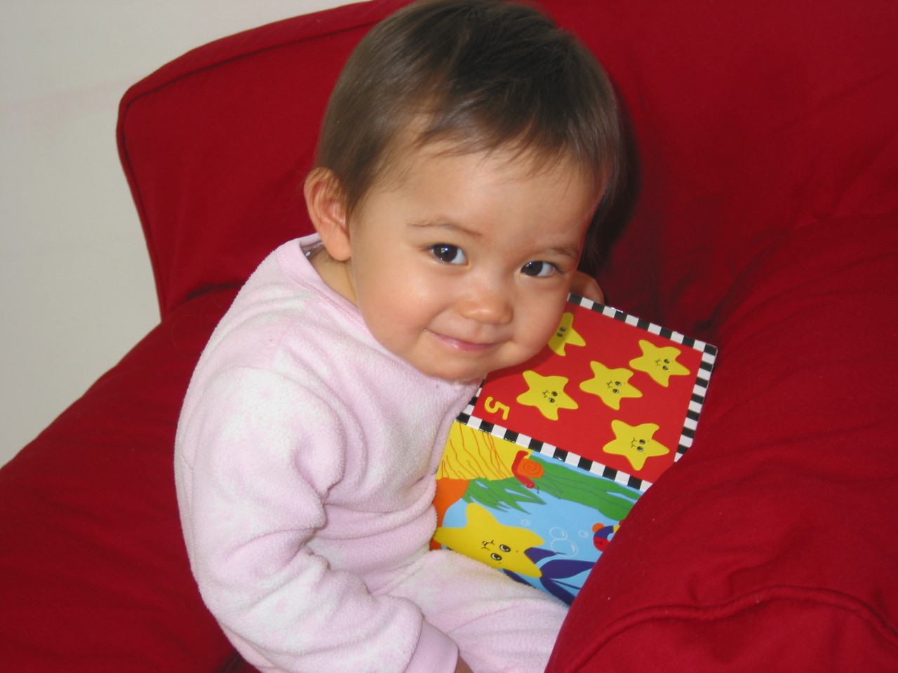 Baby Bella sits in a red beanbag chair, wearing a pink onesie and looking up from a large toy block