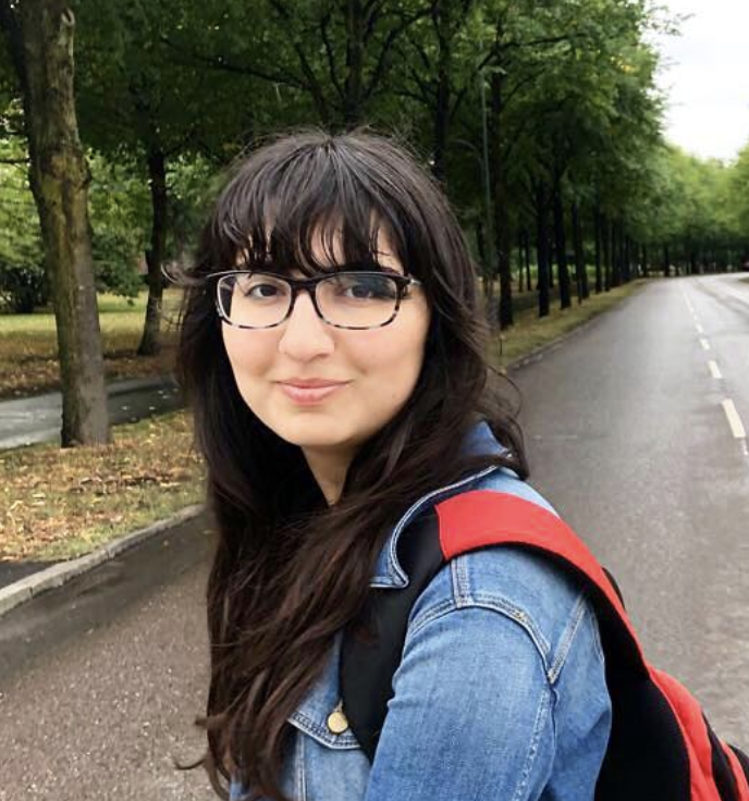 Damla wears glasses and a jean jacket while smiling on a tree lined street