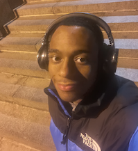 Taft stands on a staircase smiling at the camera. He has short brown hair, brown eyes and wears a black and blue coat and headphones.