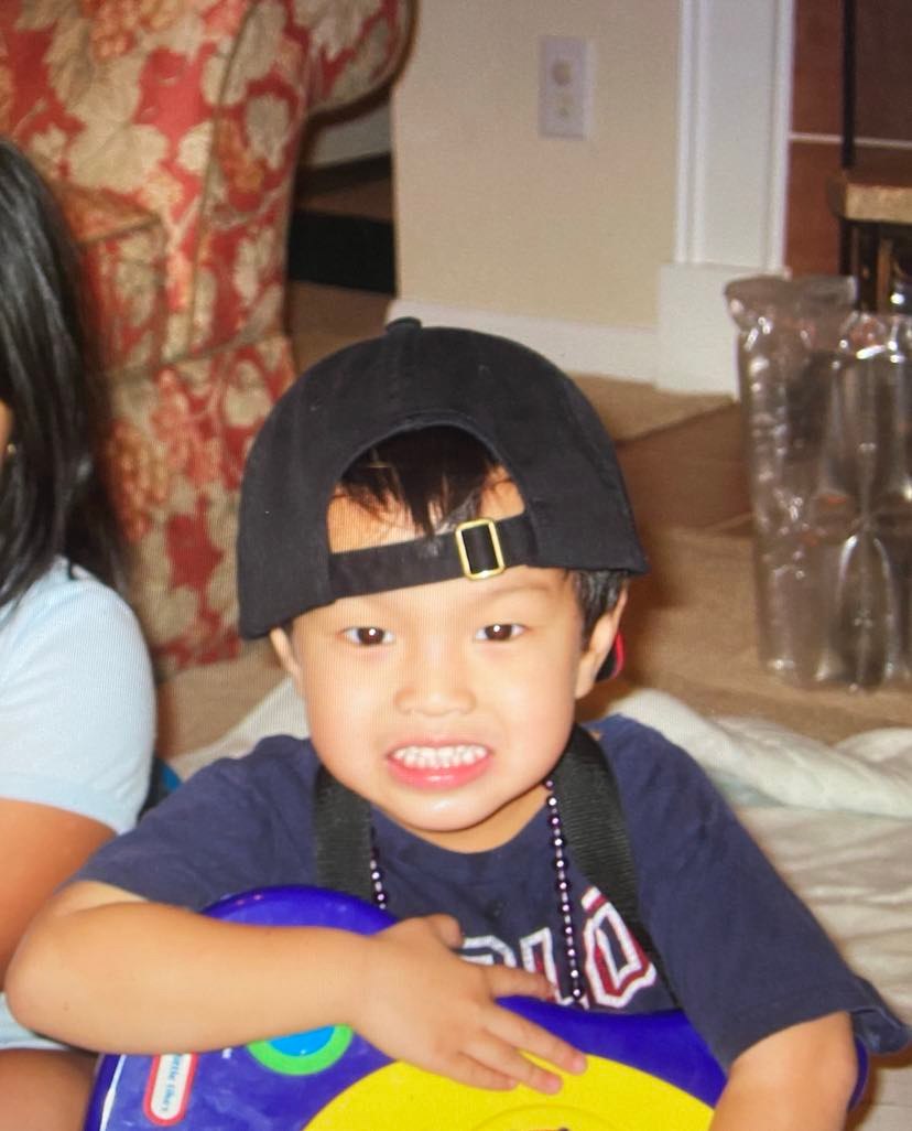 Baby George grins while wearing a backwards baseball cap, a bead necklace, and holding a toy guitar