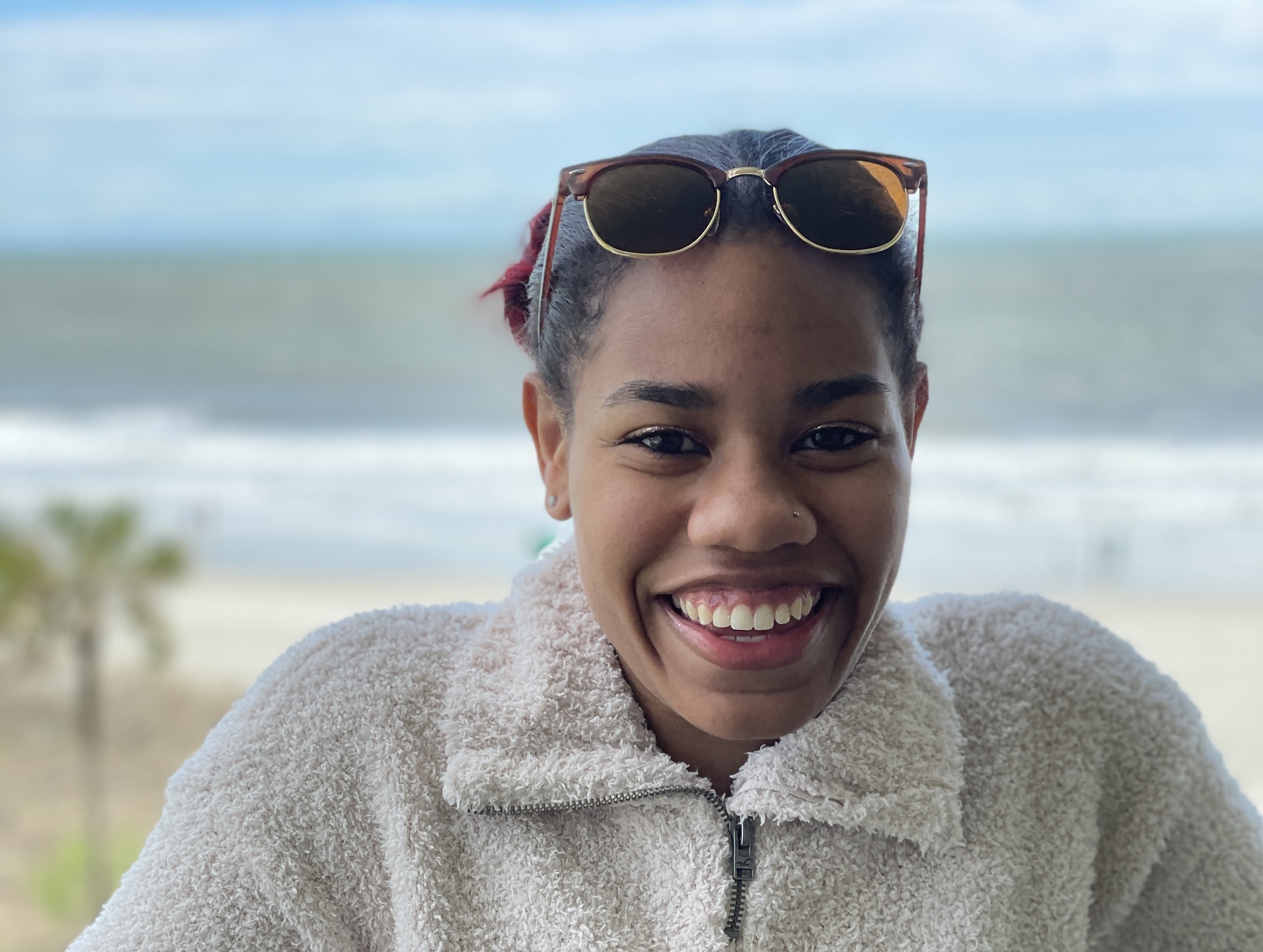 Daisja smiles in front of a beach scene. She has sunglasses on her head and wears a soft sweater.