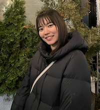 Michika stands in front of some small pine trees. She is wearing a puffy black winter coat and smiling at the camera. She has brown eyes and short dark hair with bangs.