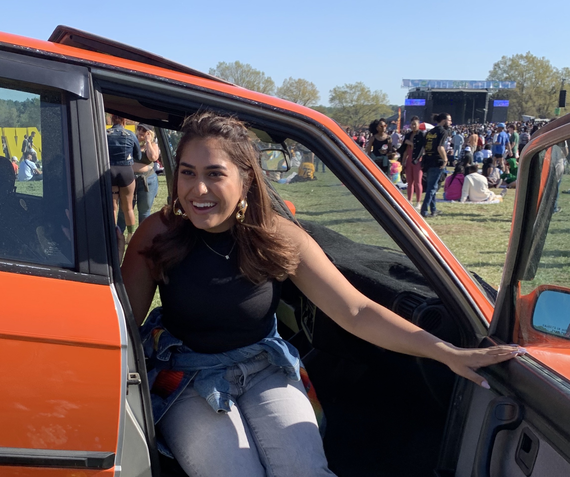 Mal sits in the passenger side of an orange retro car and smiles into the distance with flowers in the foreground and a music festival in the background