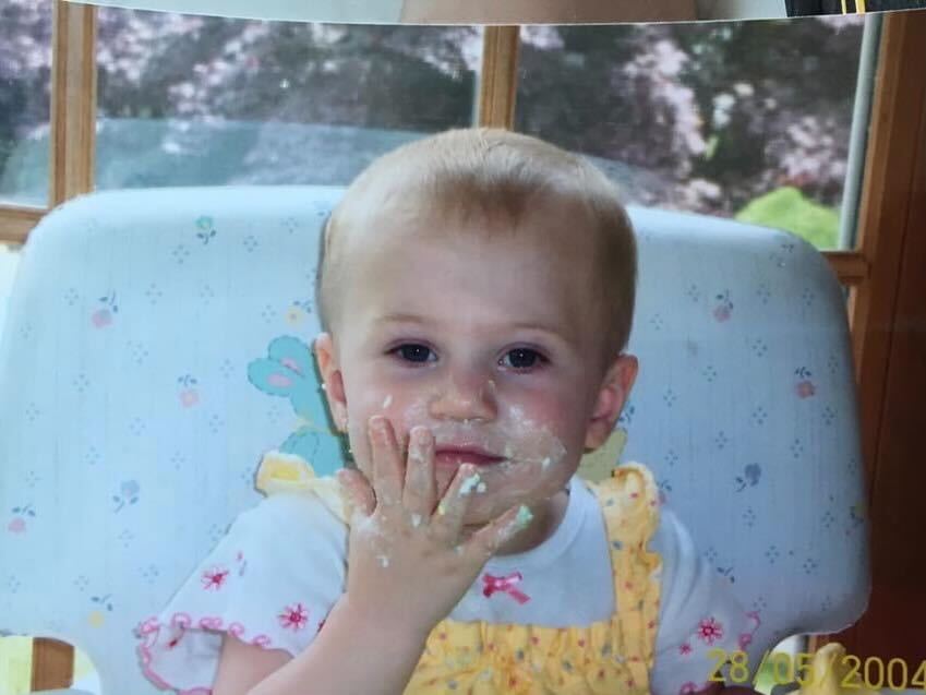 Baby Mary Kate has white food on her face and hand. She looks at the camera with a satisfied expression.