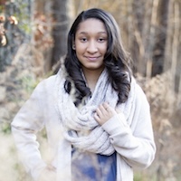  Jordan stands in the woods, smiling gently. She has shoulder length brown hair and brown eyes, and wears a cream cardigan over a blue shirt with a gray scarf.