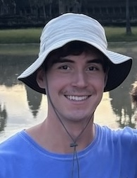 Will has brown hair and brown eyes and a kind smile. He stands in front of a lake wearing a blue shirt and floppy hat.