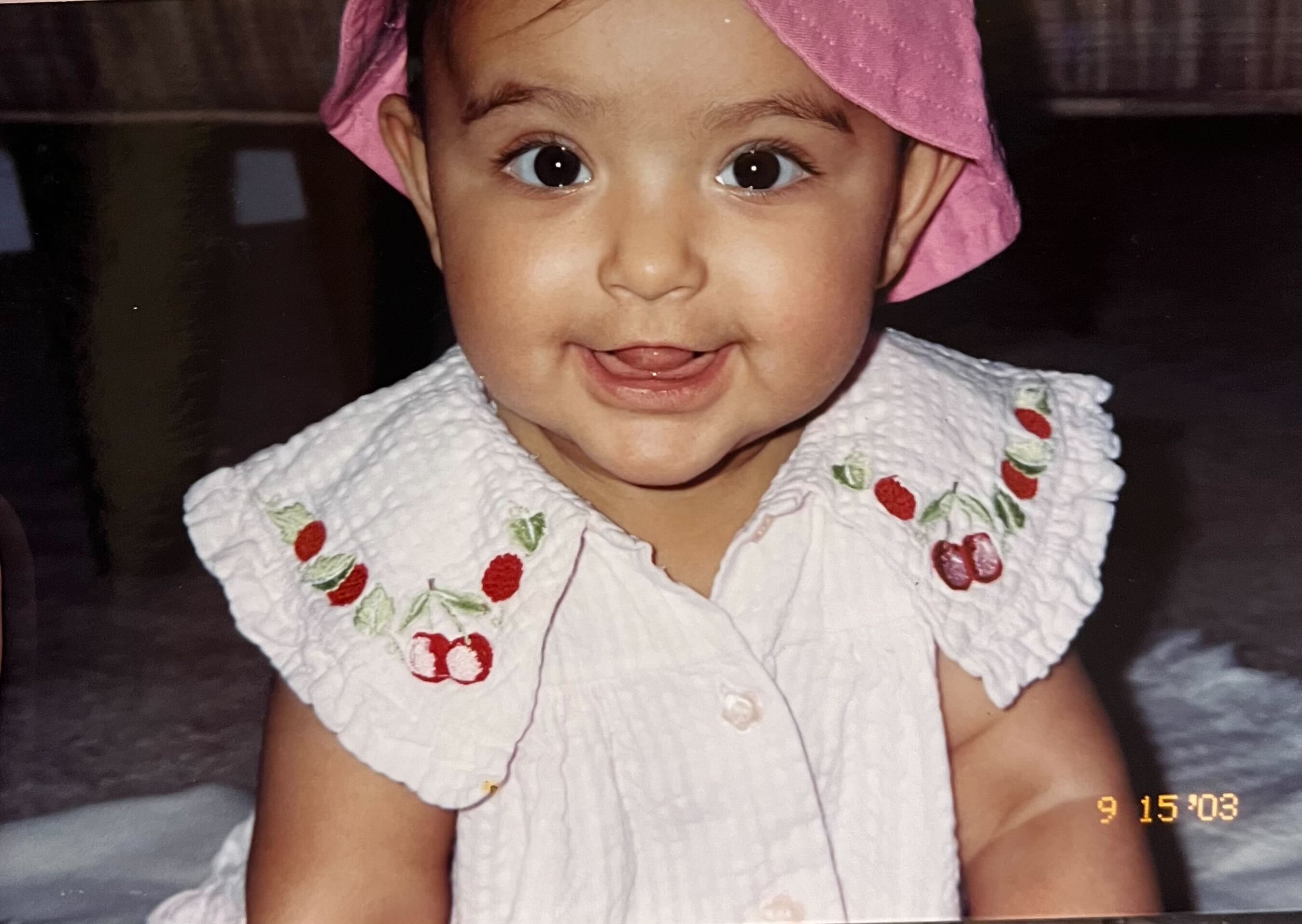Baby Serena flashes a toothless grin. She wears a white collared shirt embroidered with cherries and a pink bucket hat.