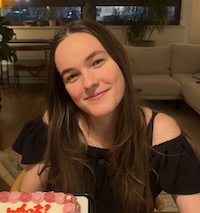 Sophie sits at a table holding a small cake and smiling. She has brown hair and brown eyes and is wearing a black top.