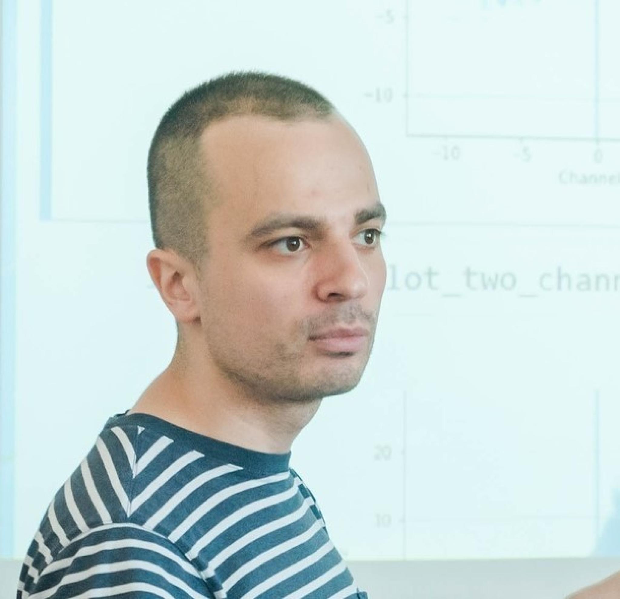 Zhenya looks to the side while standing in front of a whiteboard and wearing a blue and white striped shirt
