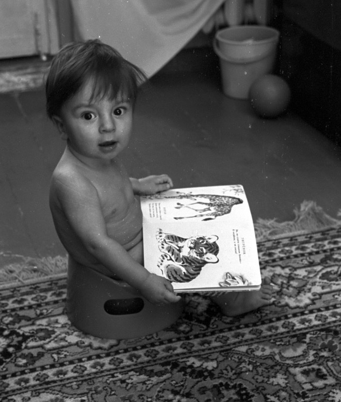 Baby Zhenya pictured in black and white, looking surprised, sitting on a booster chair and holding a picture book with tigers