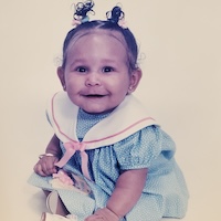 A close-up of baby Jordan. She has pig-tails in her hair and she wears a light blue dress with a white collar and a pink bow.