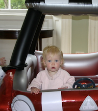 Baby Olivia wears a light pink sweater and sits behind the steering wheel of an inflatable car.