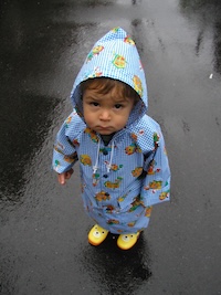 Baby Will looks up with a pout. He is wearing a blue checkered raincoat and yellow rainboots. He is not enjoying the wet and cold.