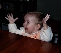 Baby Sophie wears an orange dress over a long-sleeved white shirt. She sits at a table with her arms raised above her head and mouth open like she's cheering.