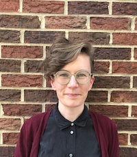 Hallie stands in front of a brick wall, wearing round glasses, a black shirt and a maroon sweater