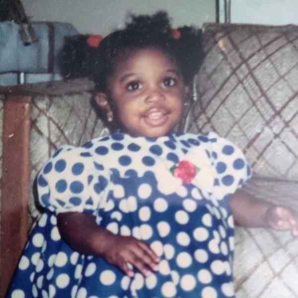 Dr. Hartman as toddler standing and grinning in front of a plaid chair in a blue polka-dotted dress