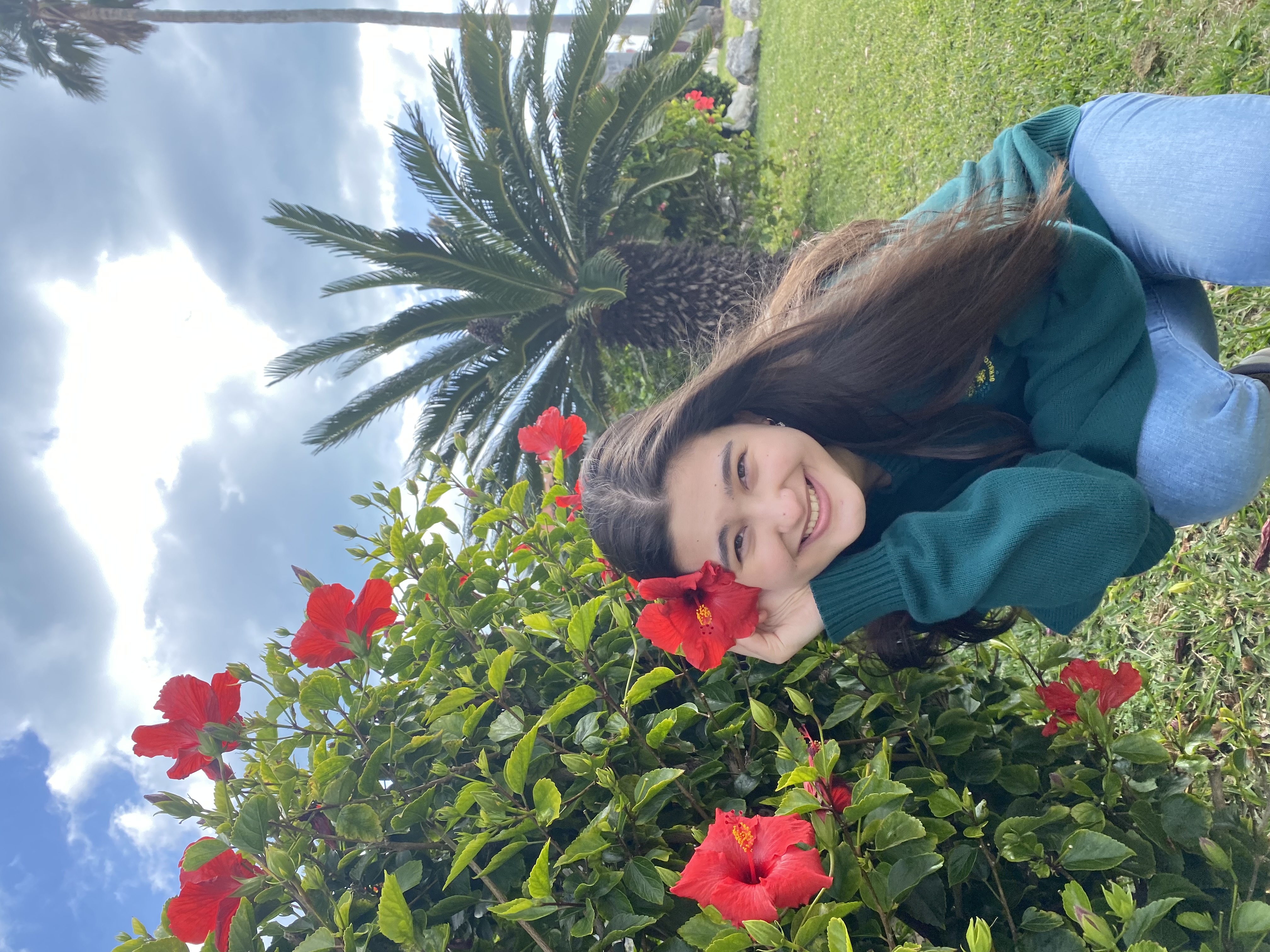 Sarah has dark brown hair and a bright smile. She kneels in front of a hibiscus bush as if putting one of the flowers in her hair, wearing a sweatshirt and jeans in a sunny tropical climate.