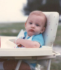 Dr. Meylan as a baby, sitting in a high chair looking bemused