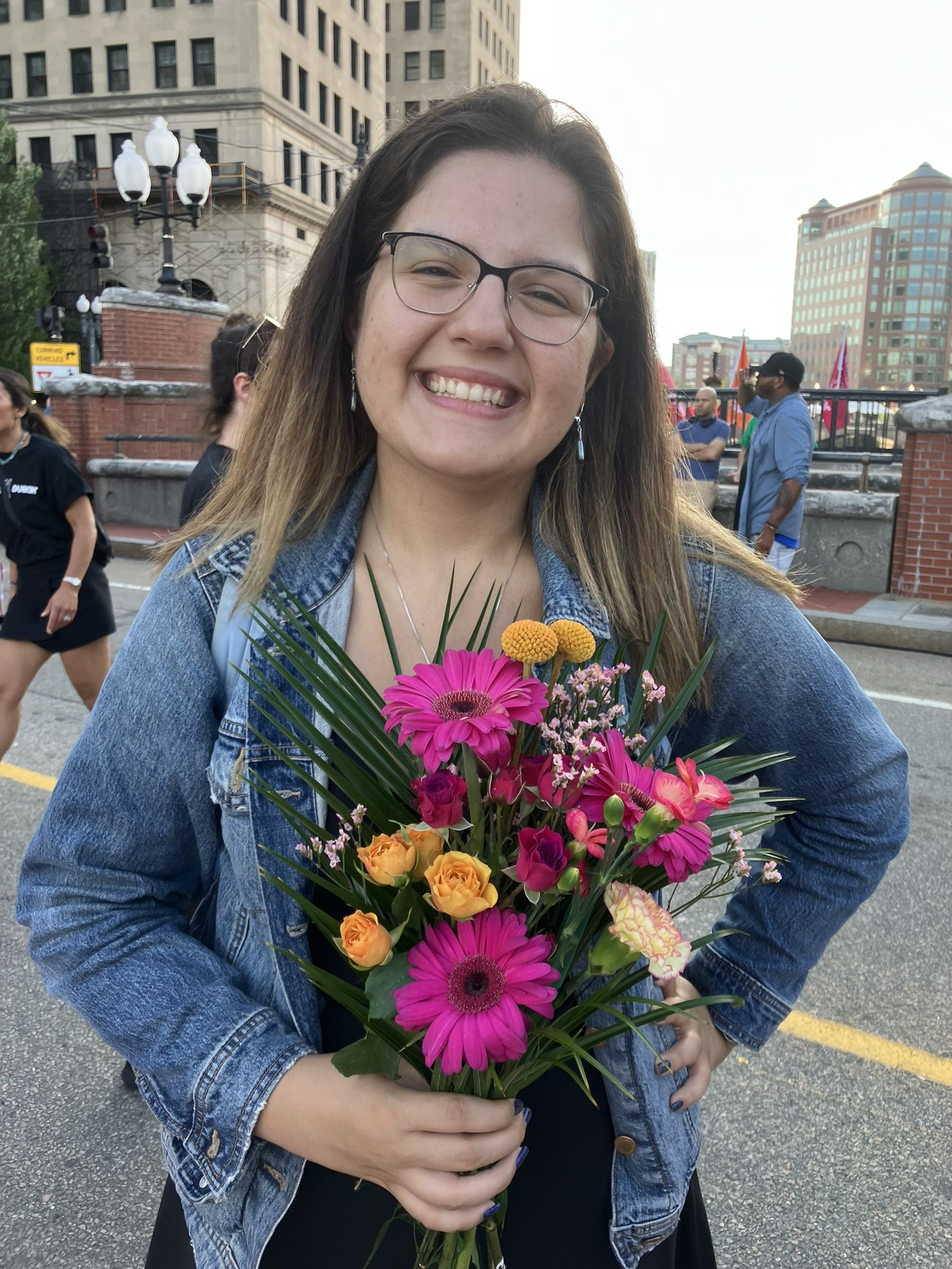 Nicole has brown hair and black square glasses. She wears a denim jacket and beams while holding a bouquet of flowers.